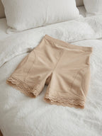 Beige shapewear shorts with lace trim at the hem, laid flat on a white bed with pillows in the background.