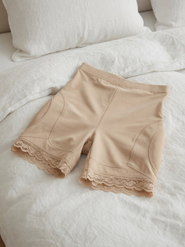 Beige shapewear shorts with lace trim at the hem, laid flat on a white bed with pillows in the background.