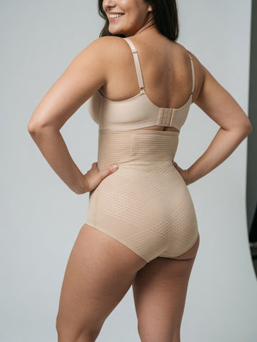 A woman shown from the back wearing a beige high-waisted compression shapewear bodysuit with a built-in bra, photographed against a light gray studio background.