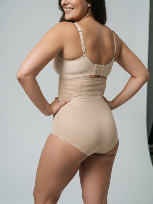 A woman shown from the back wearing a beige high-waisted compression shapewear bodysuit with a built-in bra, photographed against a light gray studio background.