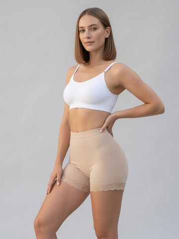 A woman modeling high-waisted beige shapewear shorts with a lace hem, paired with a white sports bra, against a solid light gray studio background.