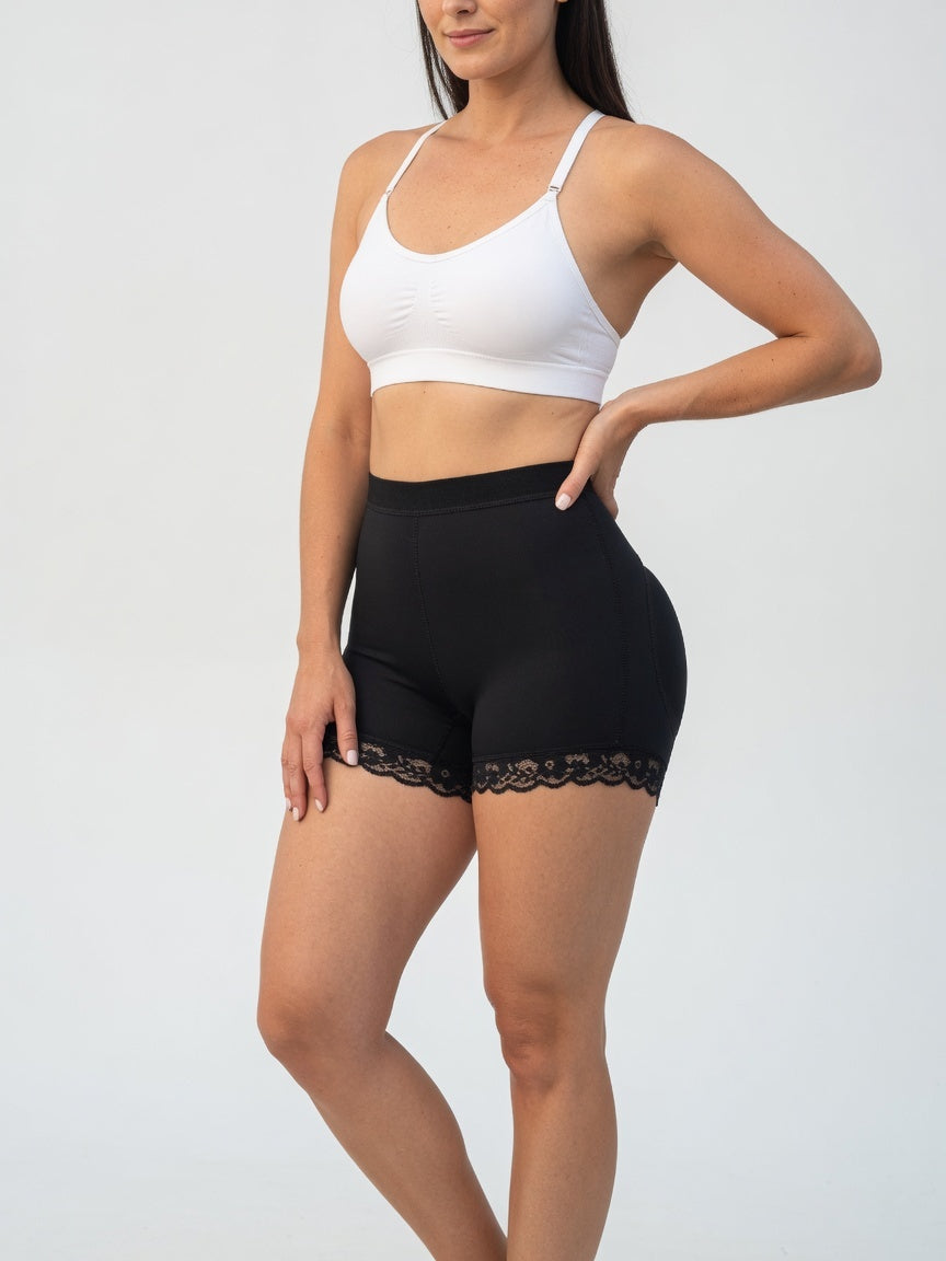 A woman modeling black high-waisted shapewear shorts with a lace hem, paired with a white sports bra, against a light gray studio background.