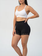A woman modeling black high-waisted shapewear shorts with a lace hem, paired with a white sports bra, against a light gray studio background.