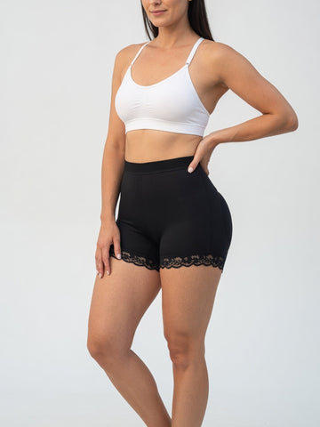 A woman modeling black high-waisted shapewear shorts with a lace hem, paired with a white sports bra, against a light gray studio background.