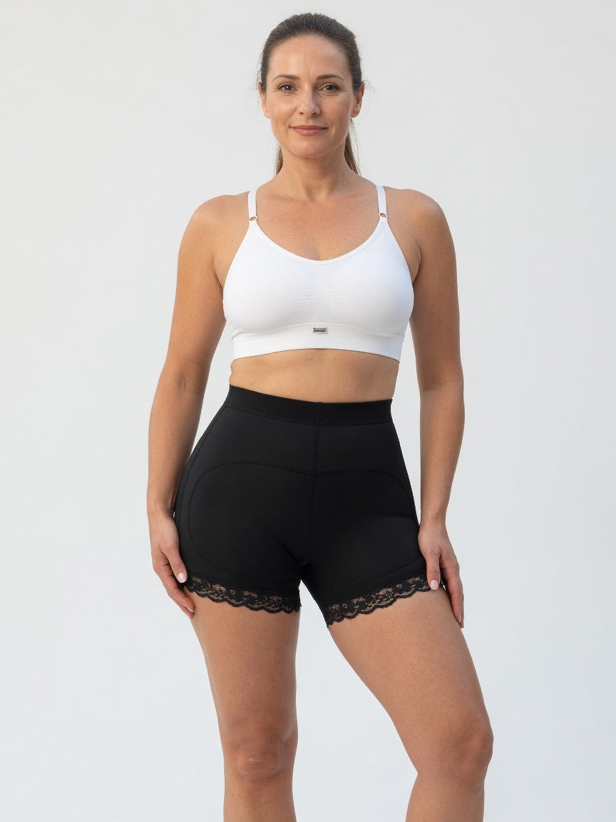 A woman modeling a white sports bra and black high-waisted athletic shorts with a lace hem, photographed against a plain light gray studio background.