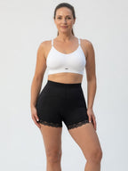 A woman modeling a white sports bra and black high-waisted athletic shorts with a lace hem, photographed against a plain light gray studio background.