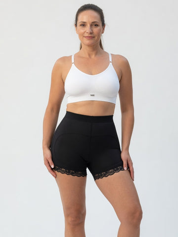 A woman modeling a white sports bra and black high-waisted athletic shorts with a lace hem, photographed against a plain light gray studio background.
