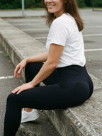 A woman sitting on a concrete curb in an outdoor parking lot, wearing a white short-sleeve t-shirt, black high-waisted leggings, and white sneakers.