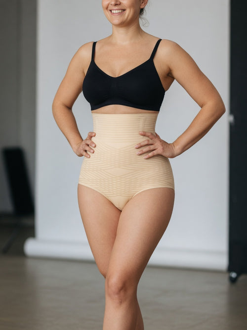 A woman modeling a high-waisted beige compression shapewear brief with a black bra, standing with hands on hips in a studio setting.