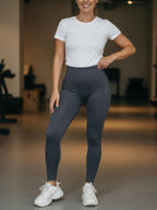 A woman modeling high-waisted dark gray athletic leggings with a white t-shirt and white sneakers in a gym setting.