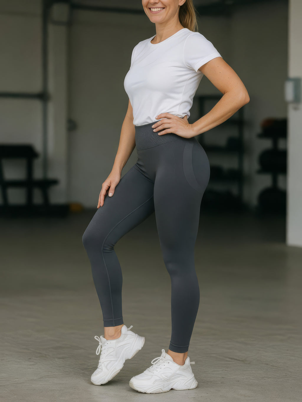 A woman modeling high-waisted dark gray leggings with a white t-shirt and white sneakers in a gym setting.