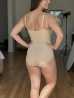 A woman wearing a beige shapewear bodysuit, shown from the back in a studio setting with a wooden floor and softbox lighting visible in the background.